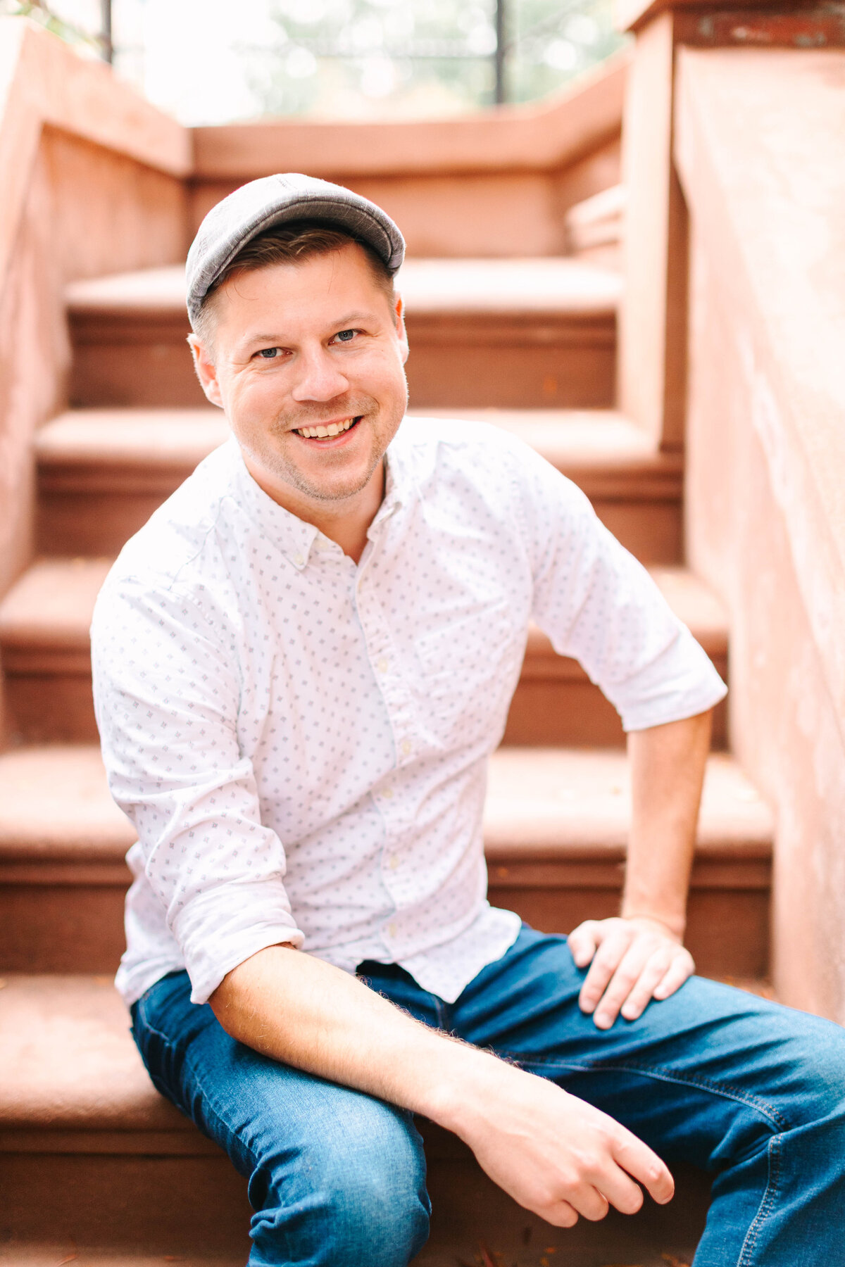 A man wearing a light-colored button-up shirt, jeans, and a gray flat cap sits on brownstone steps, smiling warmly at the camera.
