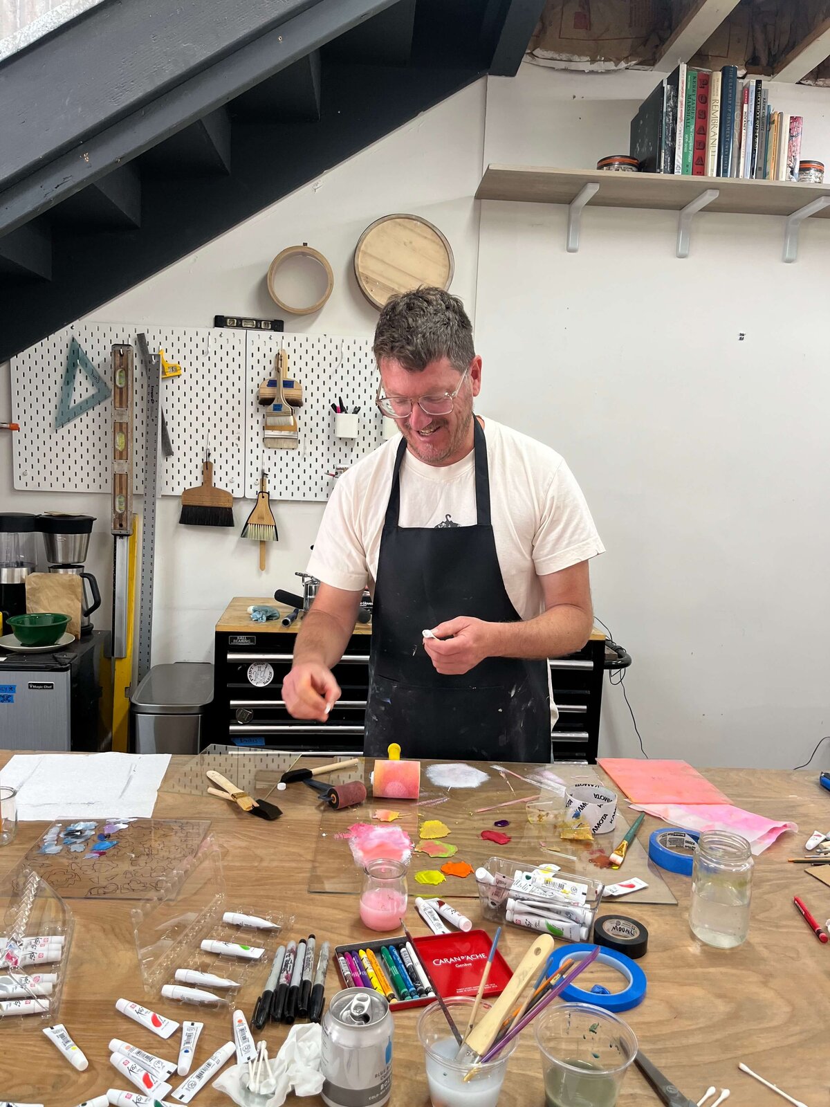 A smiling artist wearing an apron mixes pigments at a shared table, surrounded by paint tubes, rollers, and tools during the monoprinting workshop at Moonlight Press.