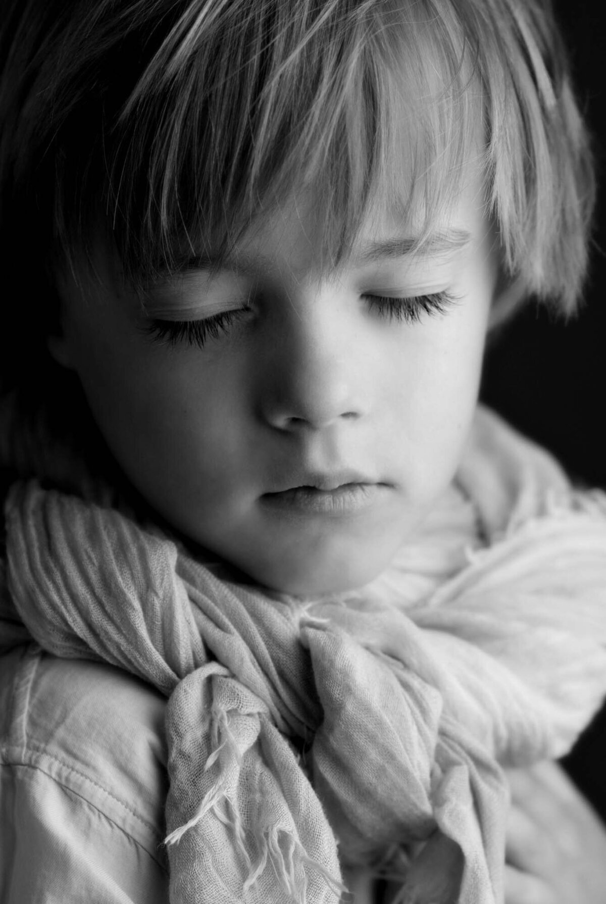 Young child with closed eyes in a black and white portrait, wearing a soft, draped scarf. The mood is serene and contemplative.