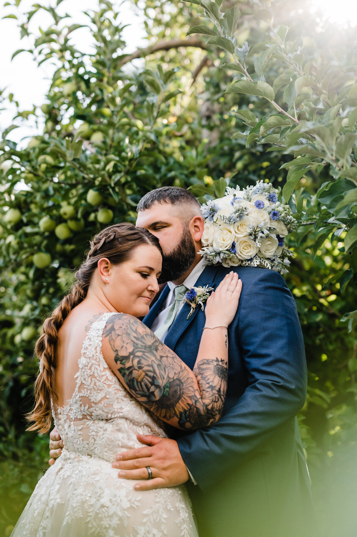bride and groom embracing in a fruit orchard while the groom kisses her head and holds her waist at showalter's orchard