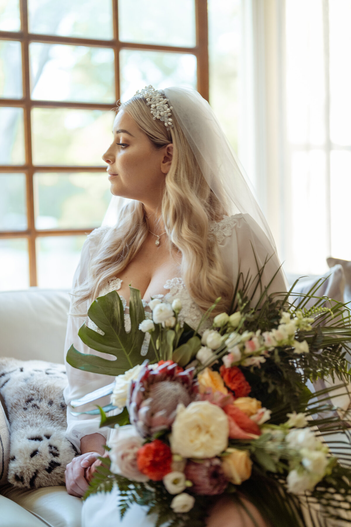Elegant portrait of a bride seated indoors on her wedding day in the DFW area. She gazes softly out of the window while holding a vibrant bouquet, showcasing her serene beauty and the intricate details of her gown and veil.