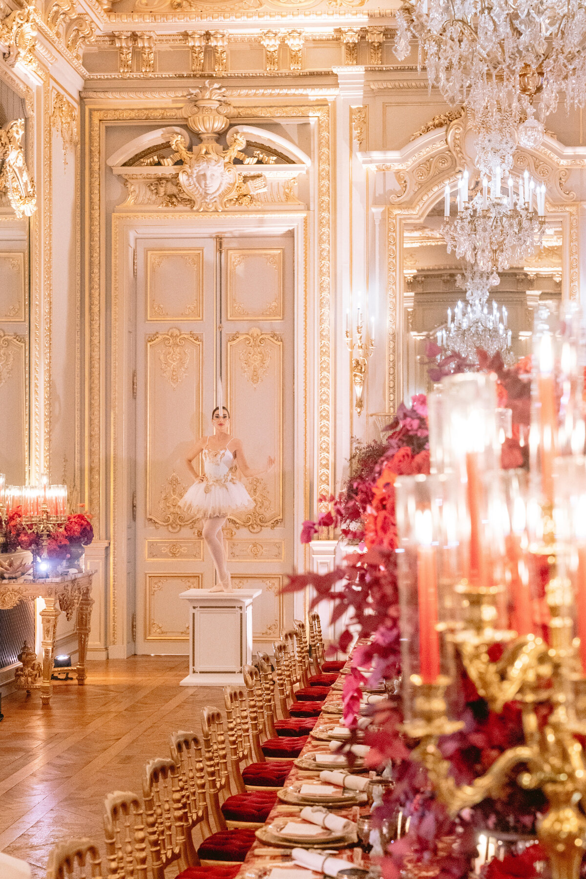 The photo was taken at the Shangri La Hotel & Ritz Paris Hotel in Paris, France by photographer Matteo Coltro in collaboration with Zen Film Works as the videographers. It features a lavish, opulent room with golden ornate decorations and chandeliers. In the center, a ballerina in a white tutu elegantly stands on a pedestal, adding a touch of grace to the luxurious setting. The scene is completed with a long, elegant dining table adorned with red flowers and candles stretching into the foreground. The table is surrounded by intricately carved chairs with red cushions, creating a truly extravagant atmosphere.