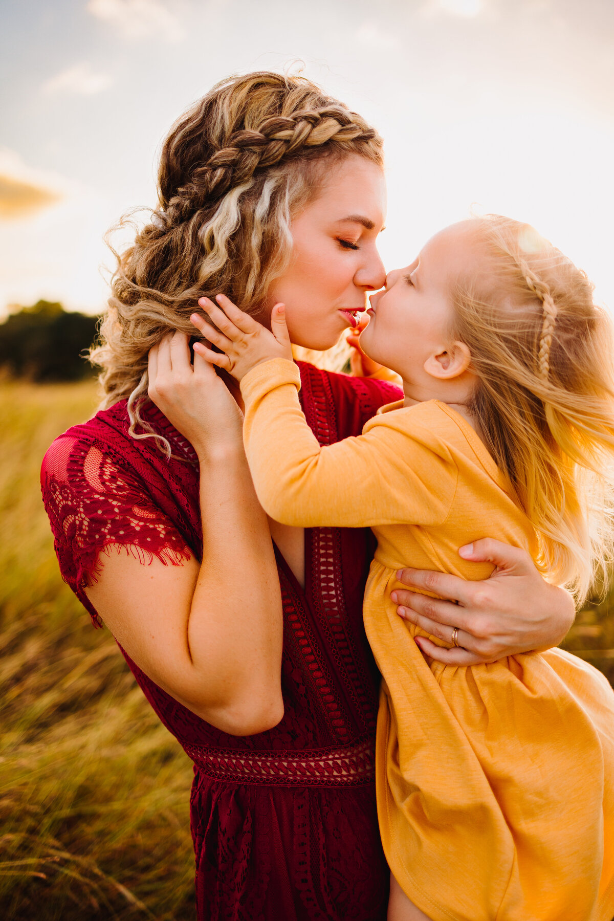 Photo of a woman with her baby girl in a garden. She has a braid in her blonde hair and with her eyes closed is giving a peck to the little girl, the woman has a red dress. The girl has a yellow dress and has a hand on the woman's cheek