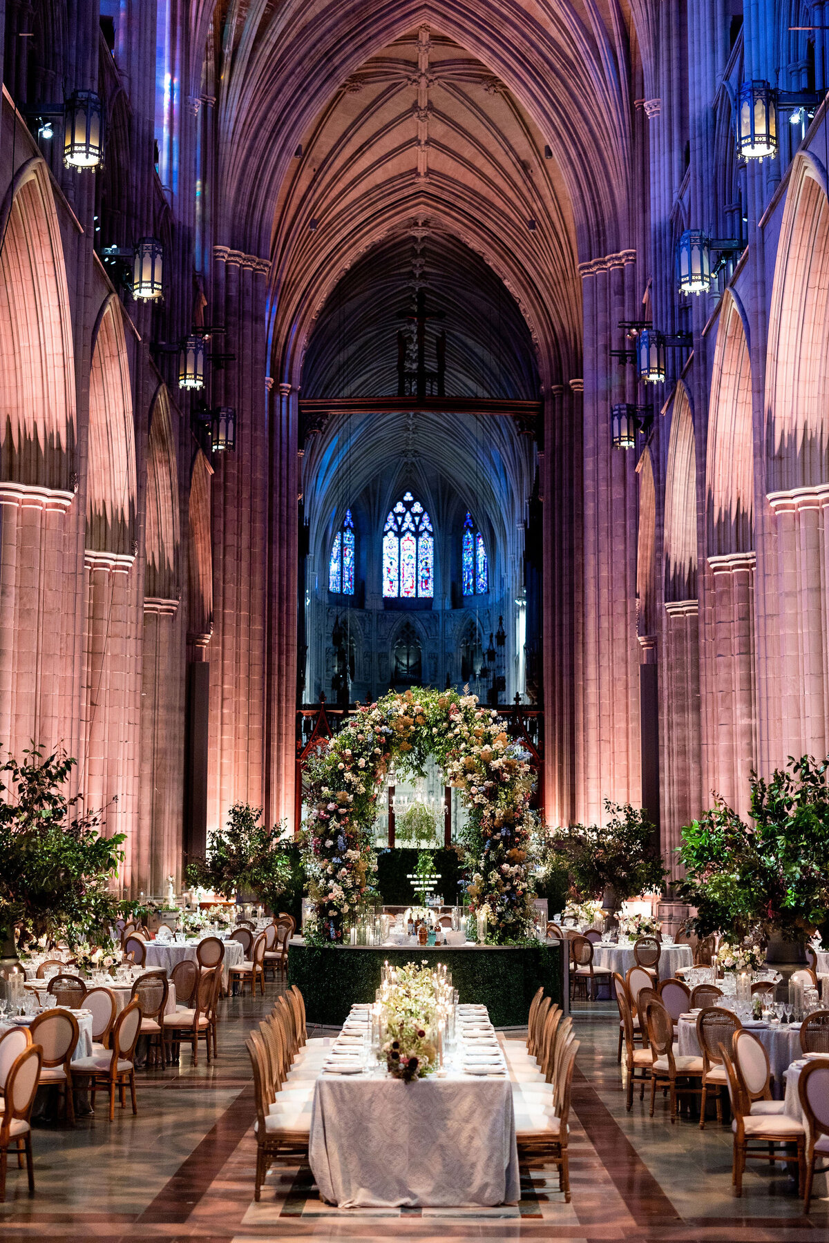 This is a stunning capture of the elegant cathedral interior at Washington National Cathedral in Washington D.C., beautifully captured by photographer Twah Dougherty. The high arched ceilings and intricate stained glass windows create a truly breathtaking atmosphere. The space is elegantly set up for a formal event, with round tables adorned in white tablecloths and delicate floral centerpieces. A grand floral archway takes center stage, gracefully leading the way to the altar, adding a touch of grandeur to the setting.