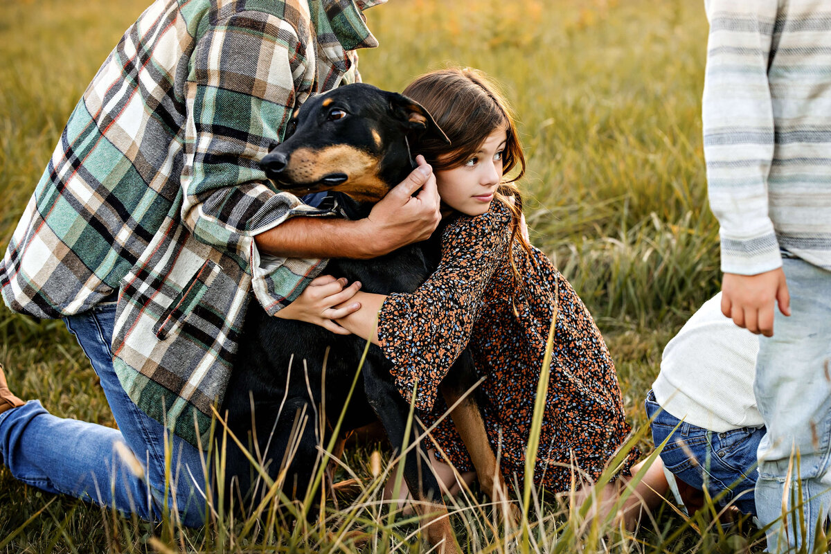 A young girl wearing a floral dress hugs a large black and brown dog in a grassy field. An adult and another child are partially visible, with the adult wearing a plaid shirt. The scene is warm and serene.