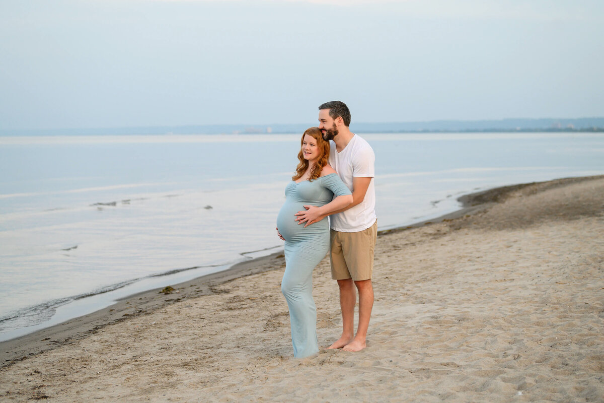 husband and wife standing in the sand looking out over the water for their beach maternity photography session in Burlington