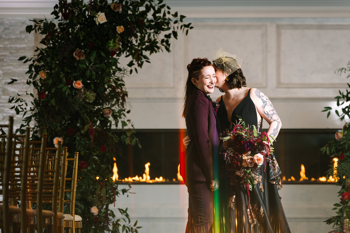 Bride in black dress kissing bride in purple suit