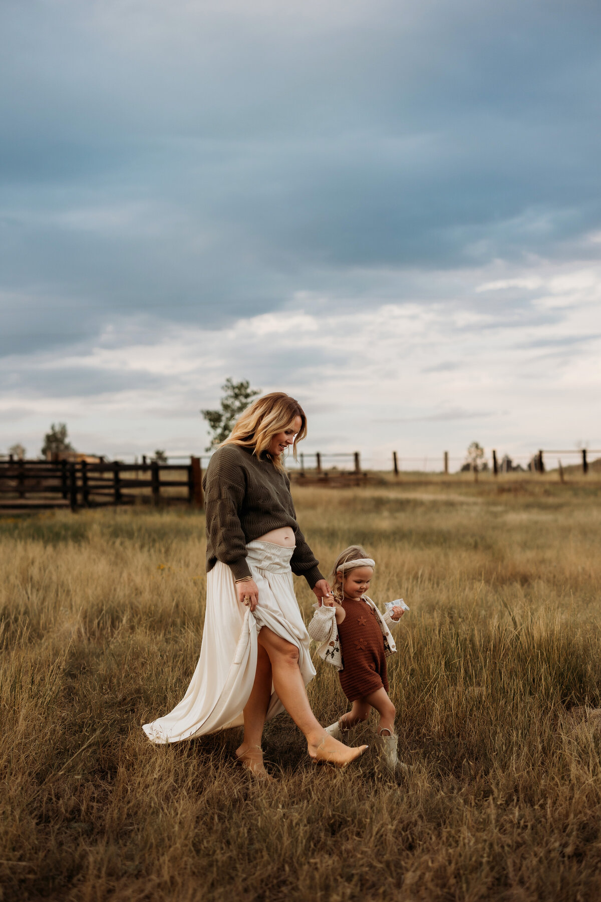 Pregnant mom and toddler daughter walking in a tall grassy field while mom's belly is exposed