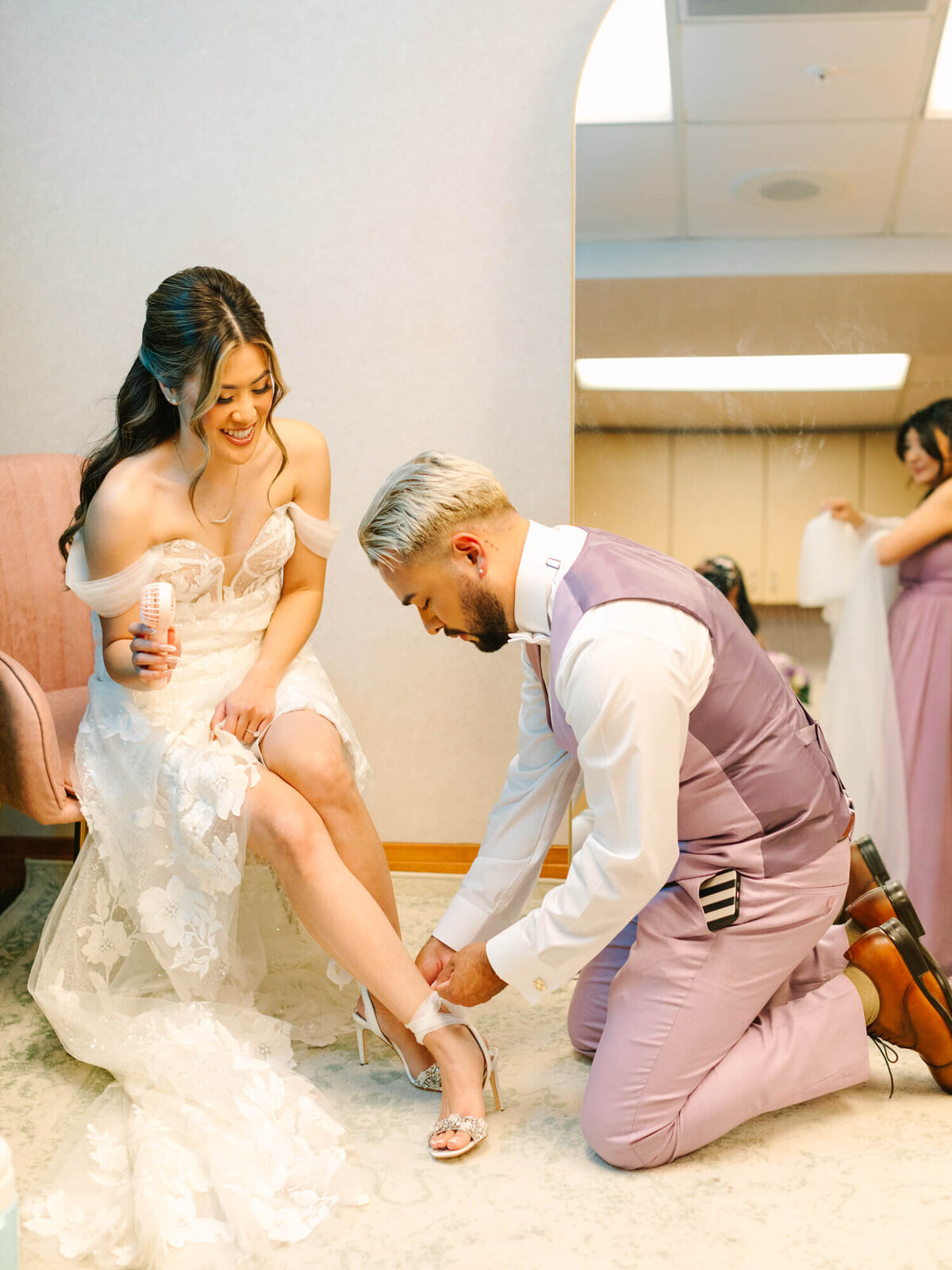 Bride in a lace gown smiles as a bridesman kneels, adjusting her sparkling high-heeled shoe. Another bridesmaid adjusts her dress in the background. Joyful, intimate moment.