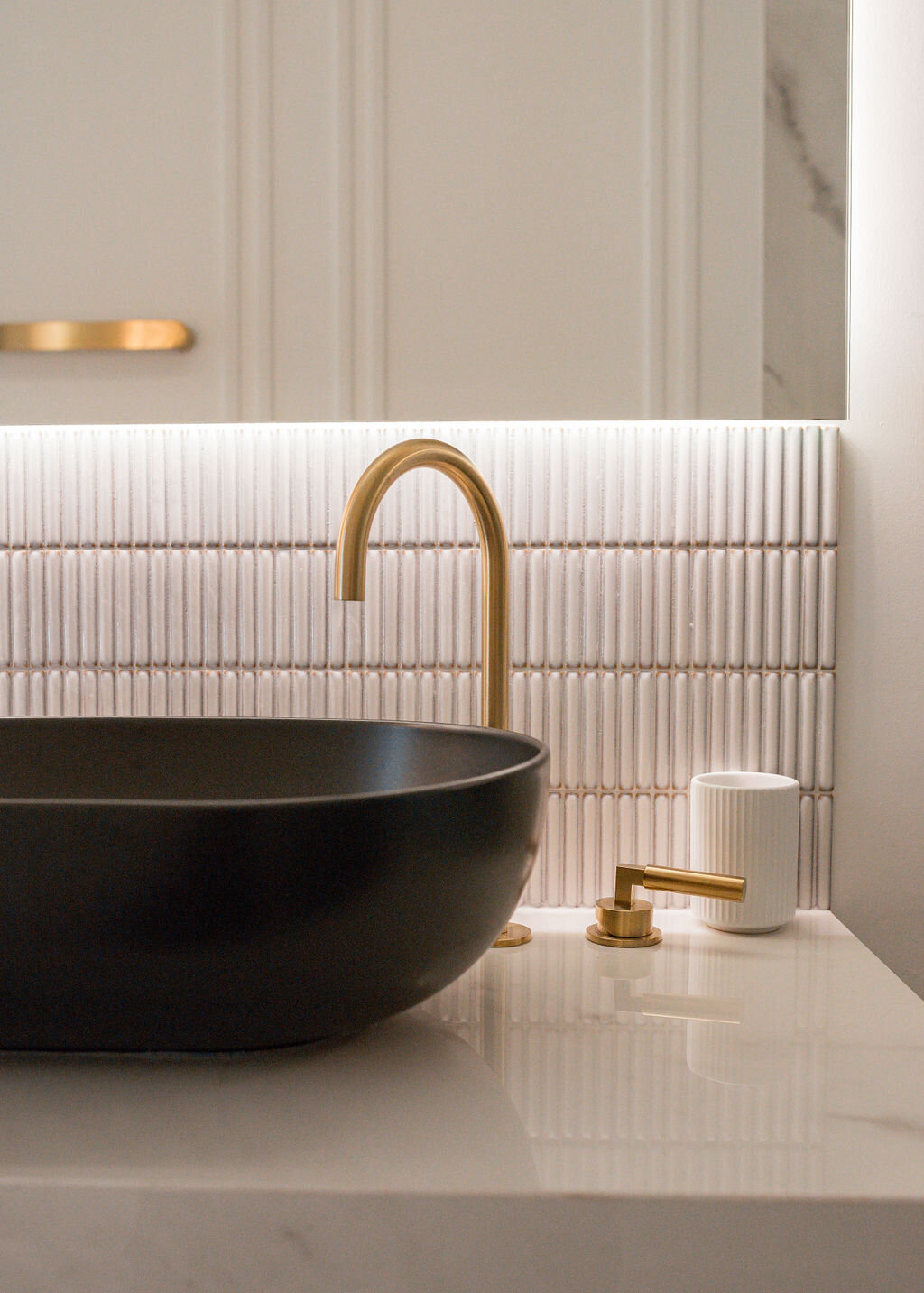 Modern bathroom with marble countertops and white tile backsplash. A black vessel sink with gold hardware sits on the counter.