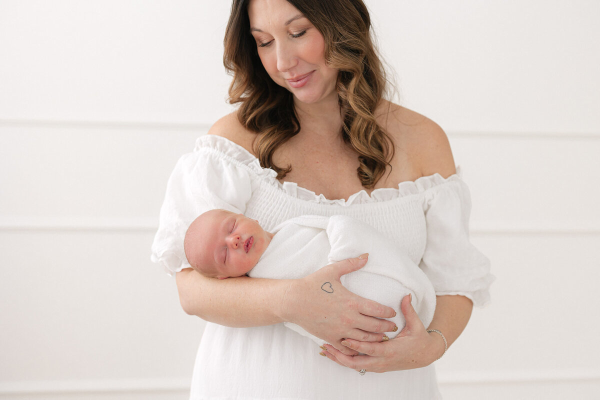 Portrait of new mom wearing a white dress while she holds her newborn baby boy swaddled in a white blanket in a bright louisville kentucky studio taken by newborn photographer missy marshall