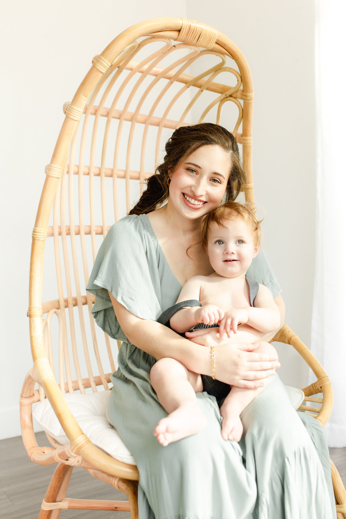 A mother wearing a sage green dress sits in a rattan chair and holds her young toddler in her arms who is wearing a green jumper in front of a white backdrop