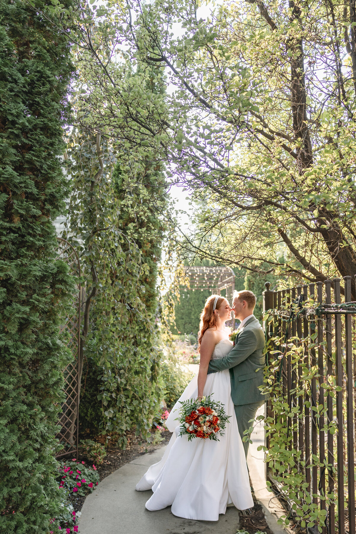 The bride and groom stand nose-to-nose under a canopy of green vines and trees, sharing an intimate moment while holding a bouquet of orange and white roses.
