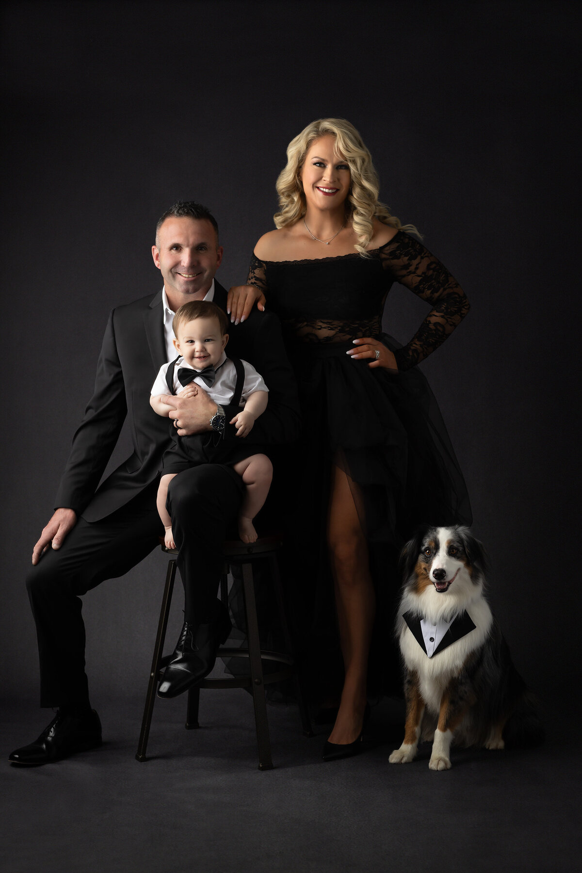 Elegant family of three and their dog dressed in formal black attire posing against a dark studio backdrop for a Dallas milestone photographer session.