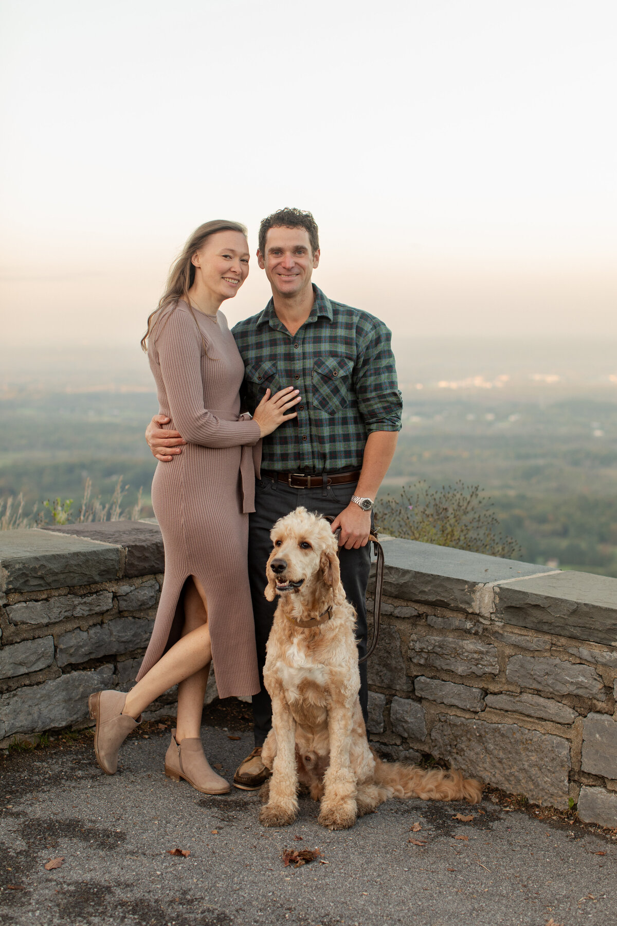 Engaged couple smile at the camera, their dog sitting on the ground in front of them