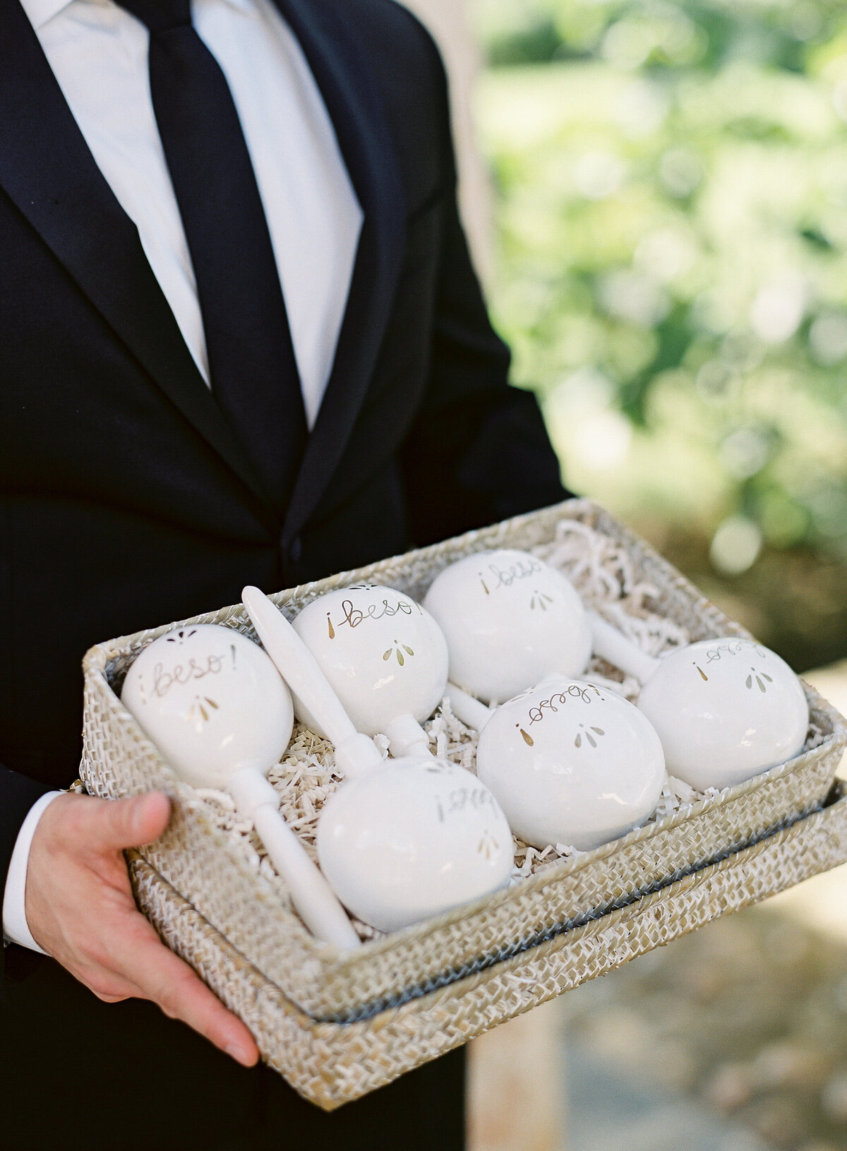 A man in a black suit and tie holds a rectangular woven basket filled with white maracas. Each maraca displays words like best and love in gold cursive. The background showcases lush greenery, creating a tropical ambiance suitable for a Caribbean destination wedding at one of Dominican Republic's top venues. This image was beautifully captured by photographer Laura Gordon.