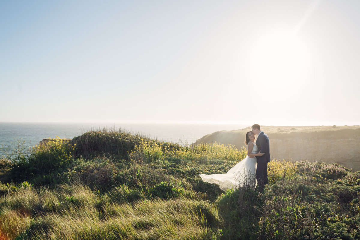Bride and groom on meadow cliffs of Shark Fin Cove Santa Cruz holding each other close while the sunlight grazes the grass