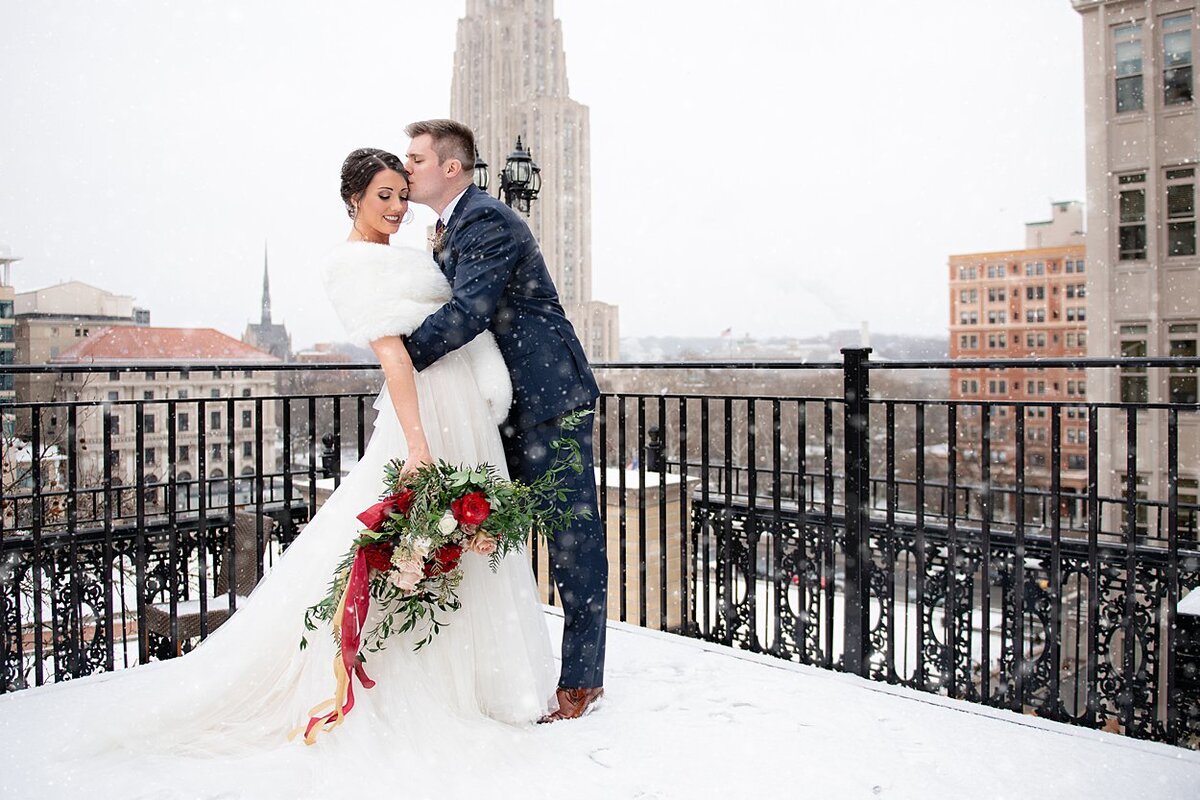 Groom kissing Bride in fur wrap on forehead on snowy rooftop in Pittsburgh, PA with Cathedral of Learning in background