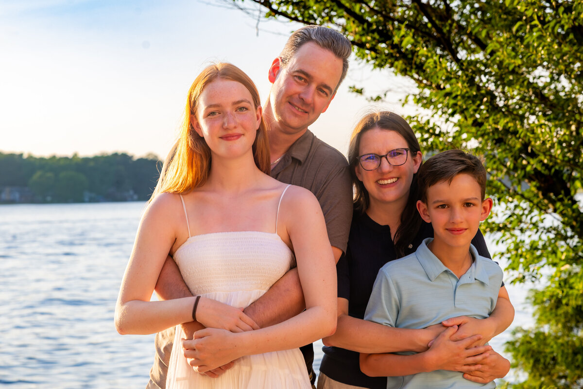 family of four posing snuggly under a tree in front of a lake, captured by Capture Joy Photography, Northern Virginia family photographer.
