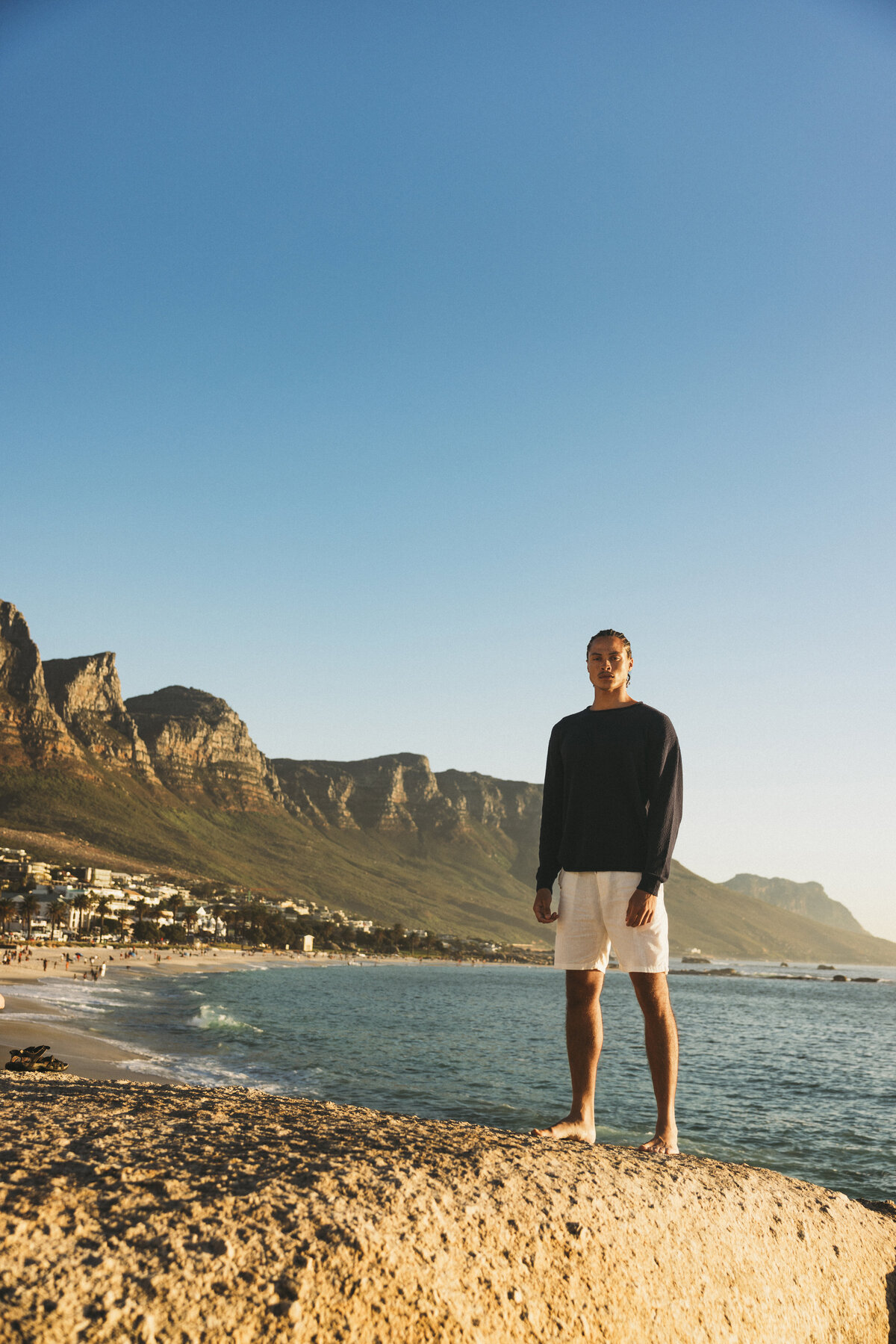 Male model Timothe standing barefoot on a rock at the beach in Cape Town, wearing a dark sweater and white shorts, with the Twelve Apostles mountain range and the ocean in the background.