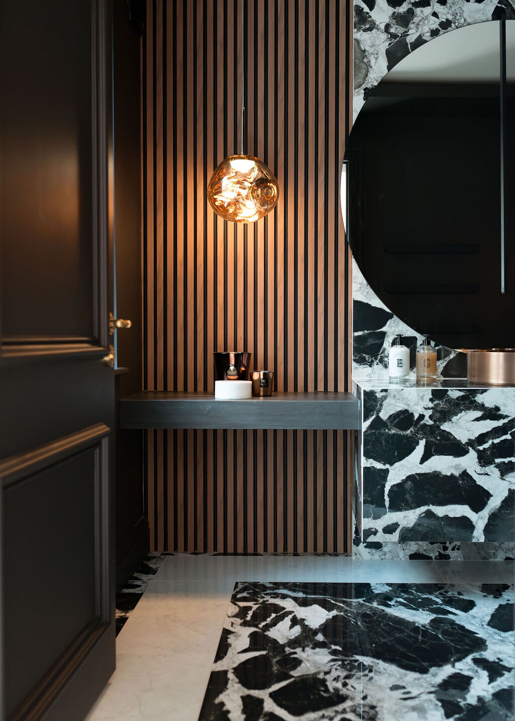 A modern bathroom with a large round mirror, black and white marble sink and countertop, wooden slatted wall, and pendant light fixture.