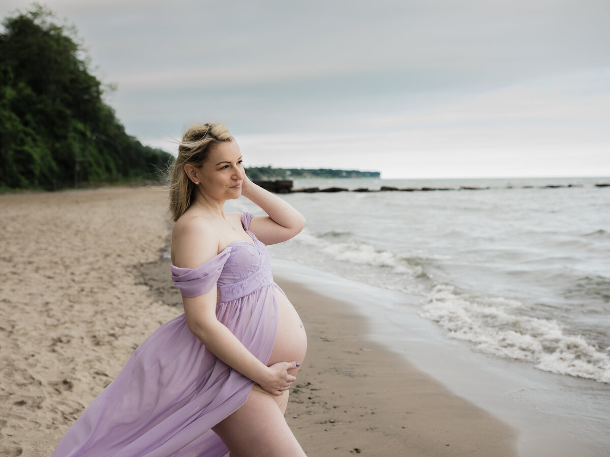 pregnant mother in purple maternity gown holding baby bump on beach for maternity photoshoot 