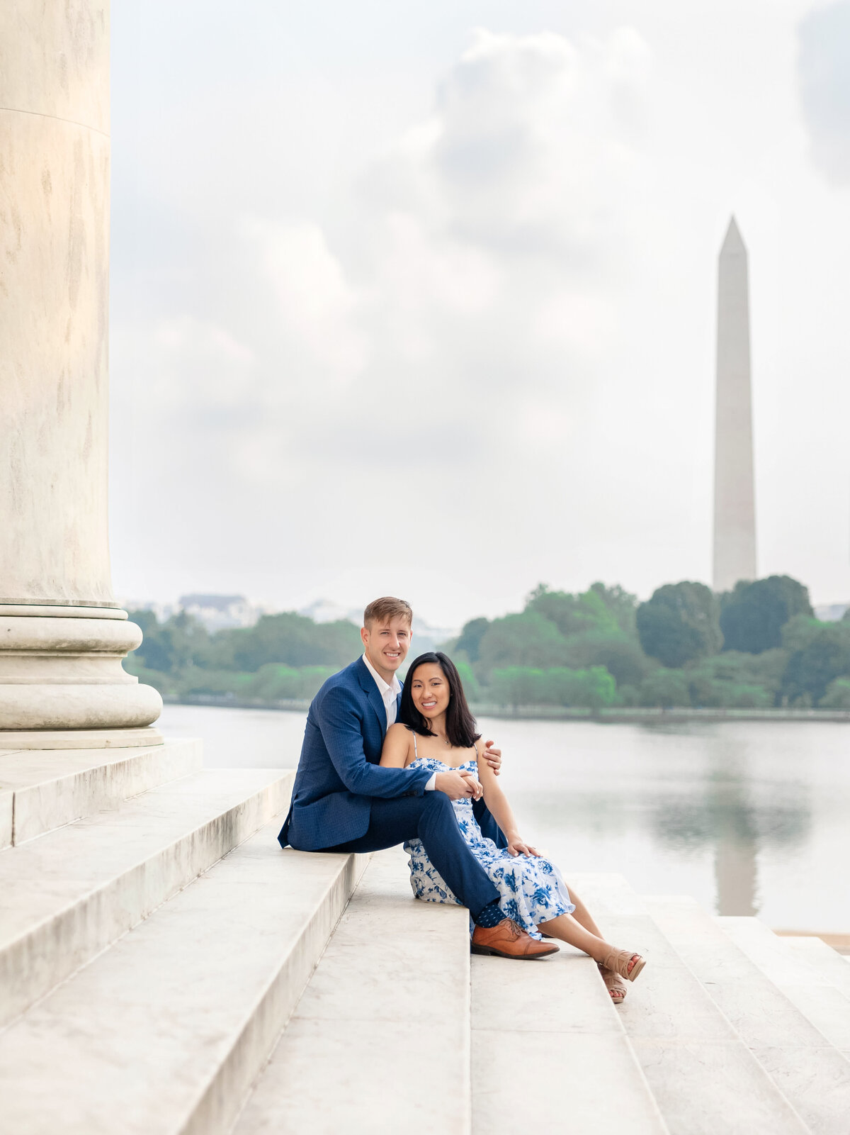 A couple sits on marble steps, embracing and smiling. The Washington Monument is visible in the background across the water. The sky is partly cloudy, and the scene conveys a peaceful, romantic atmosphere.