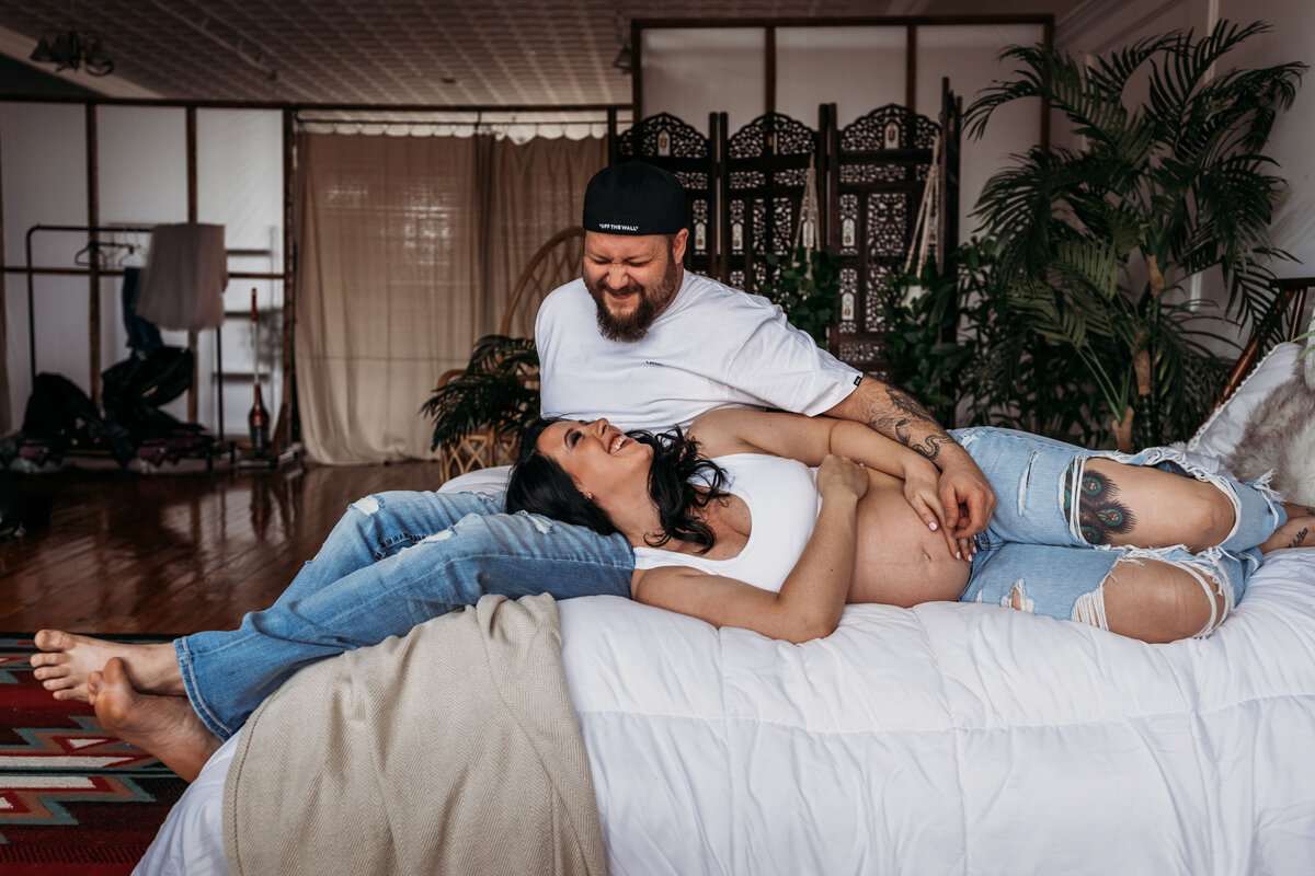 A pregnant couple with white tees and wripped jeans laughing and laying in bed