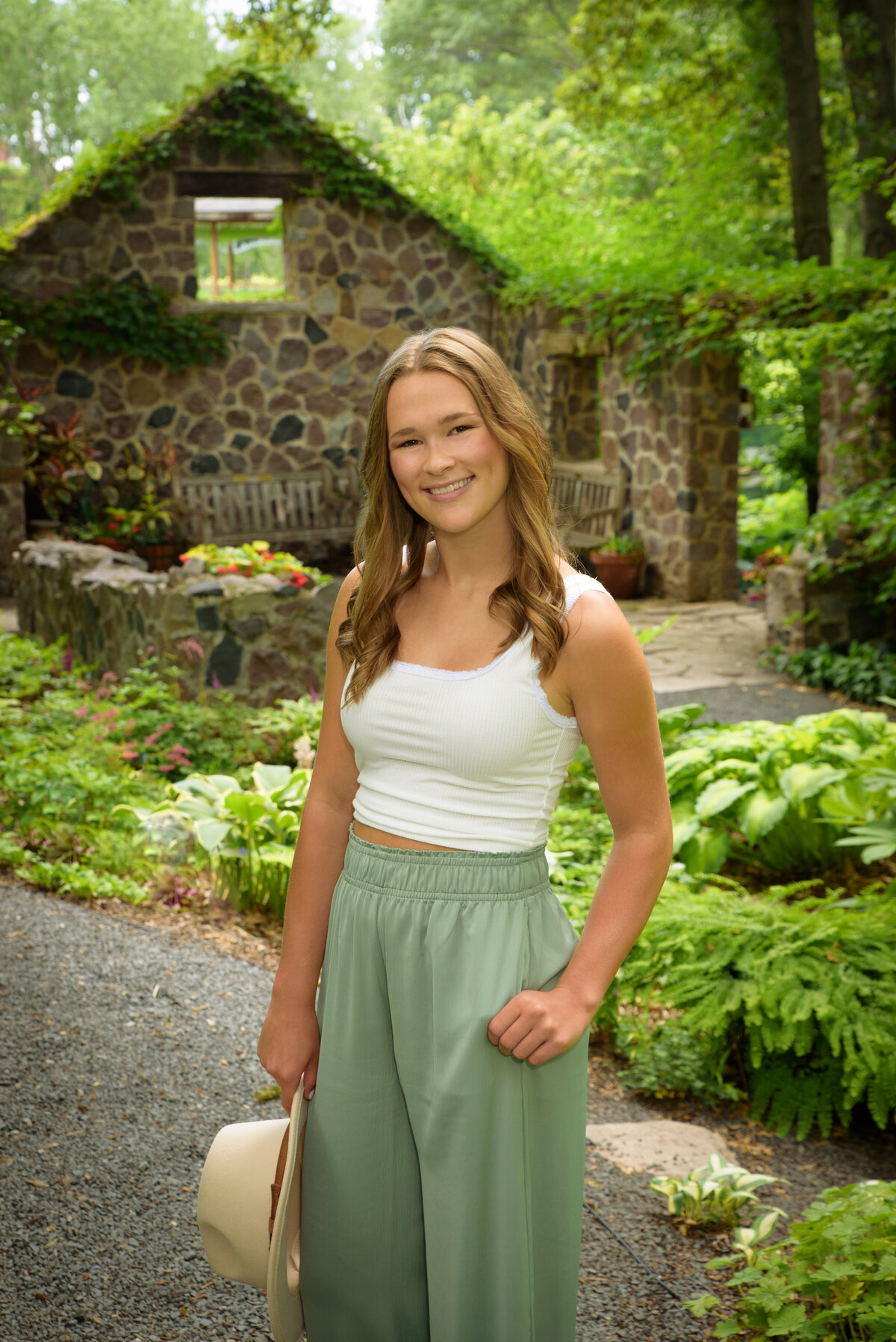 Luxemburg Casco High School senior girl wearing long sage green pants and a cream hat sitting by creek in the Hosta garden at the Green Bay Botanical Gardens in Green Bay, Wisconsin