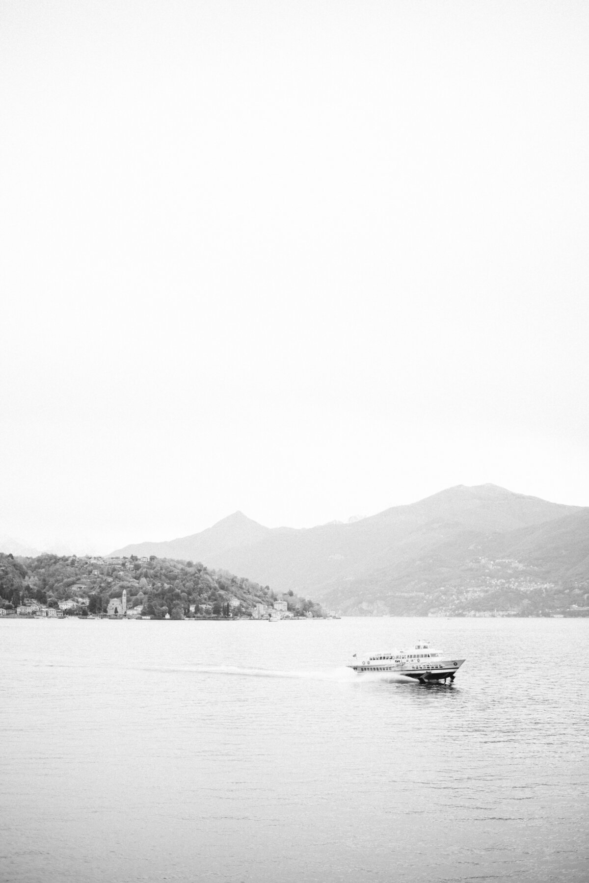 Minimalist black and white photo of a small boat gliding across Lake Como, framed by misty mountains and a quiet shoreline in the distance.