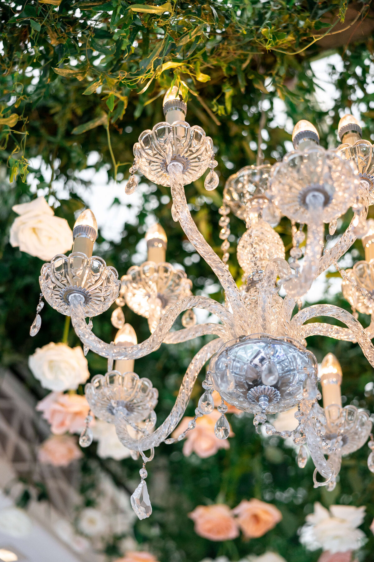 A crystal chandelier with lit bulbs hangs from a ceiling adorned with green foliage and white and pink roses, creating an elegant and romantic atmosphere.