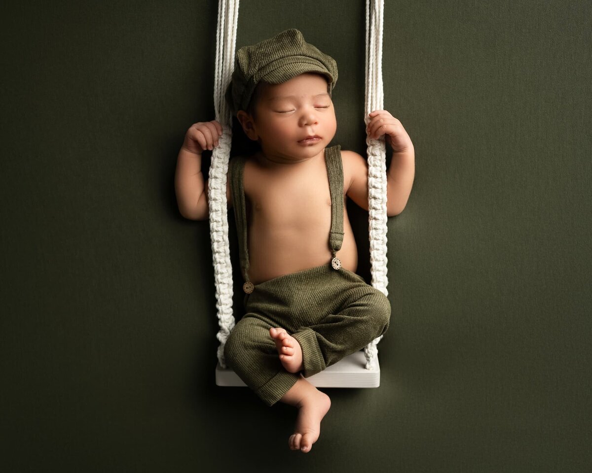 newborn boy wearing green corduroy overalls and matching pageboy hat sitting on a white swing