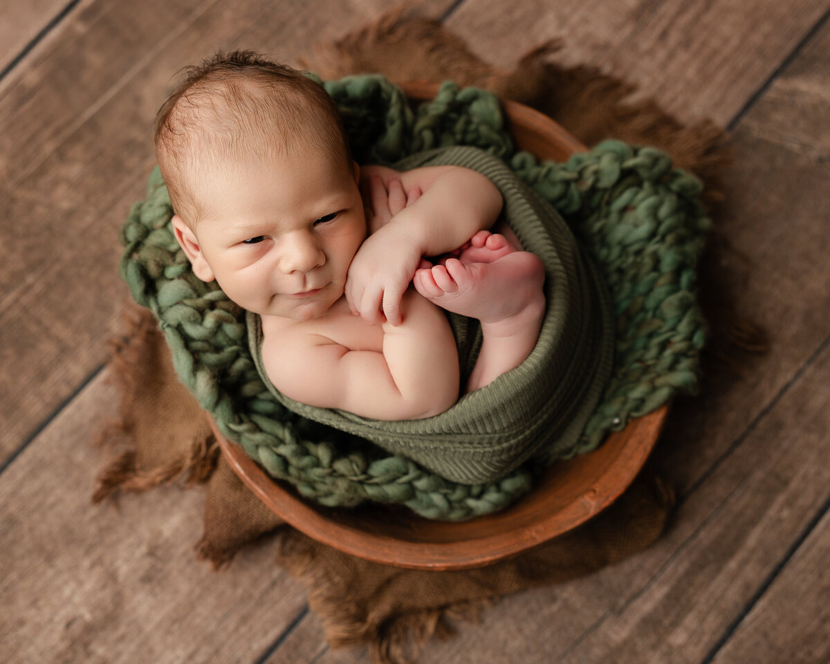 Newborn baby boy wrapped in earthy green tones, posed in a wooden bowl on a brown textured backdrop during his studio session. Captured by Inland Empire newborn photographer Kaitlyn Dawn Photography.