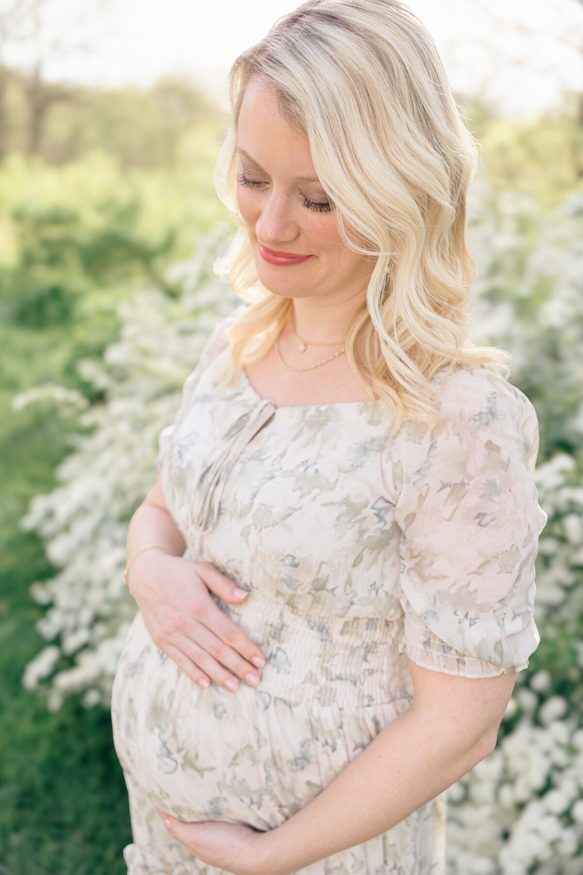 A pregnant mother in a sage green floral dress looks down lovingly at her baby belly during her session at Newfields with Indianapolis maternity photographer, Brittney Lear.