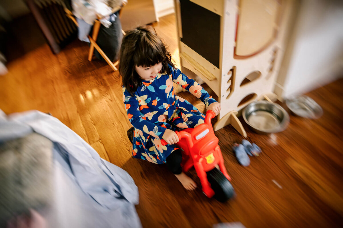 4 year old girl races through a messy living room floor on her toy red motorcycle.