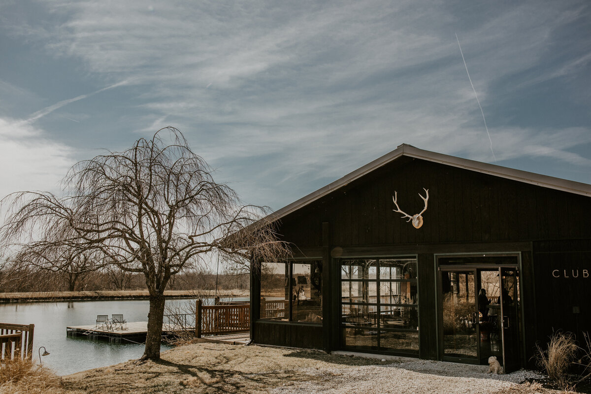 Dark wooden clubhouse with a deck besides a pond with a bare tree and white antlers upfront.