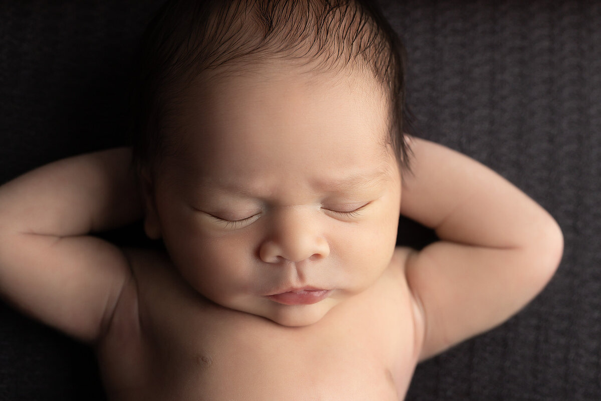 Calm newborn baby posed with hands behind head on a dark gray backdrop by best Dallas newborn photographer.