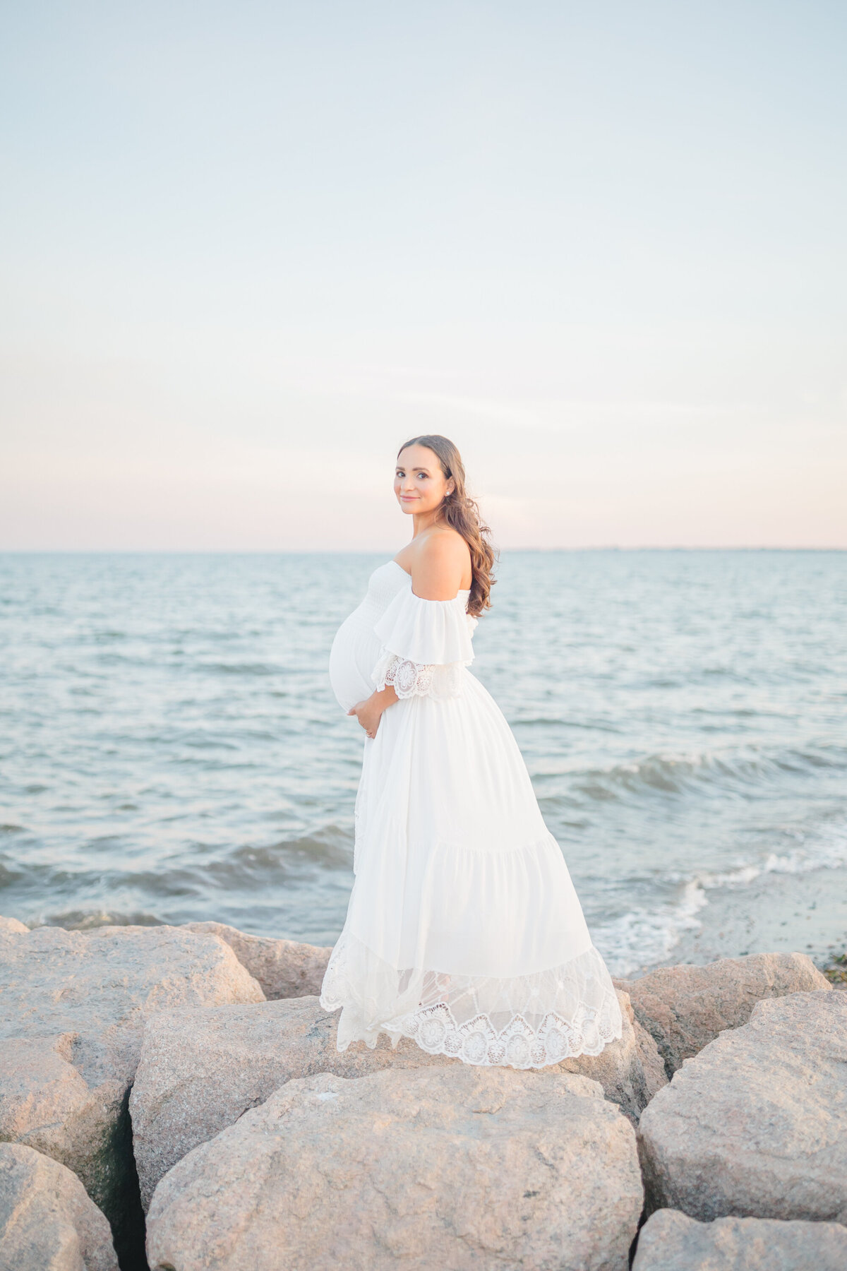 Pregnant mom standing on a cliffside at sunset, wearing a flowing white gown, with soft golden light surrounding her