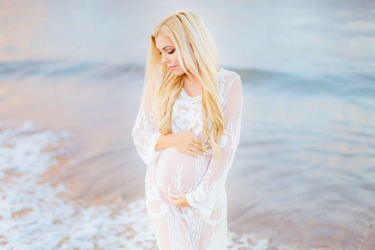 Blonde woman wearing long sleeved white maternity dress glances over her right shoulder and holds her baby belly during her Wailea photography session