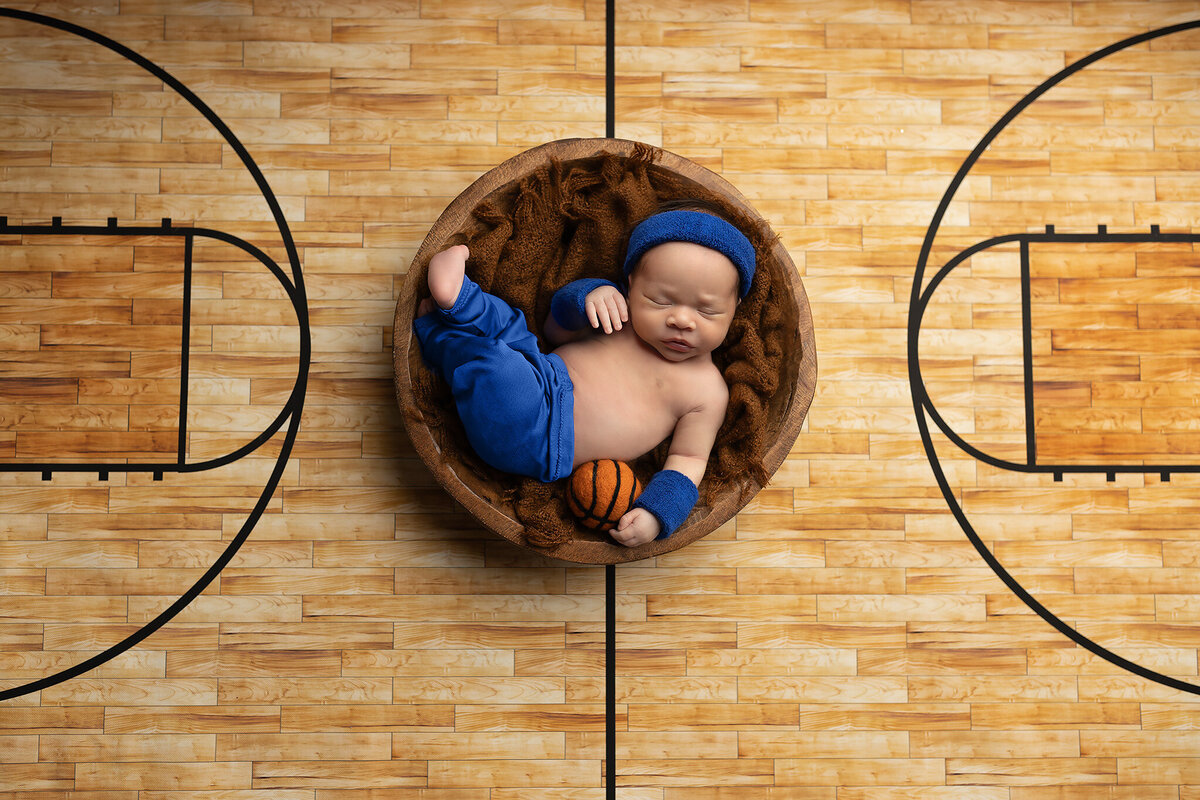 Newborn baby dressed in blue sports outfit with a mini basketball, posed on a wood court backdrop – Southlake newborn photography