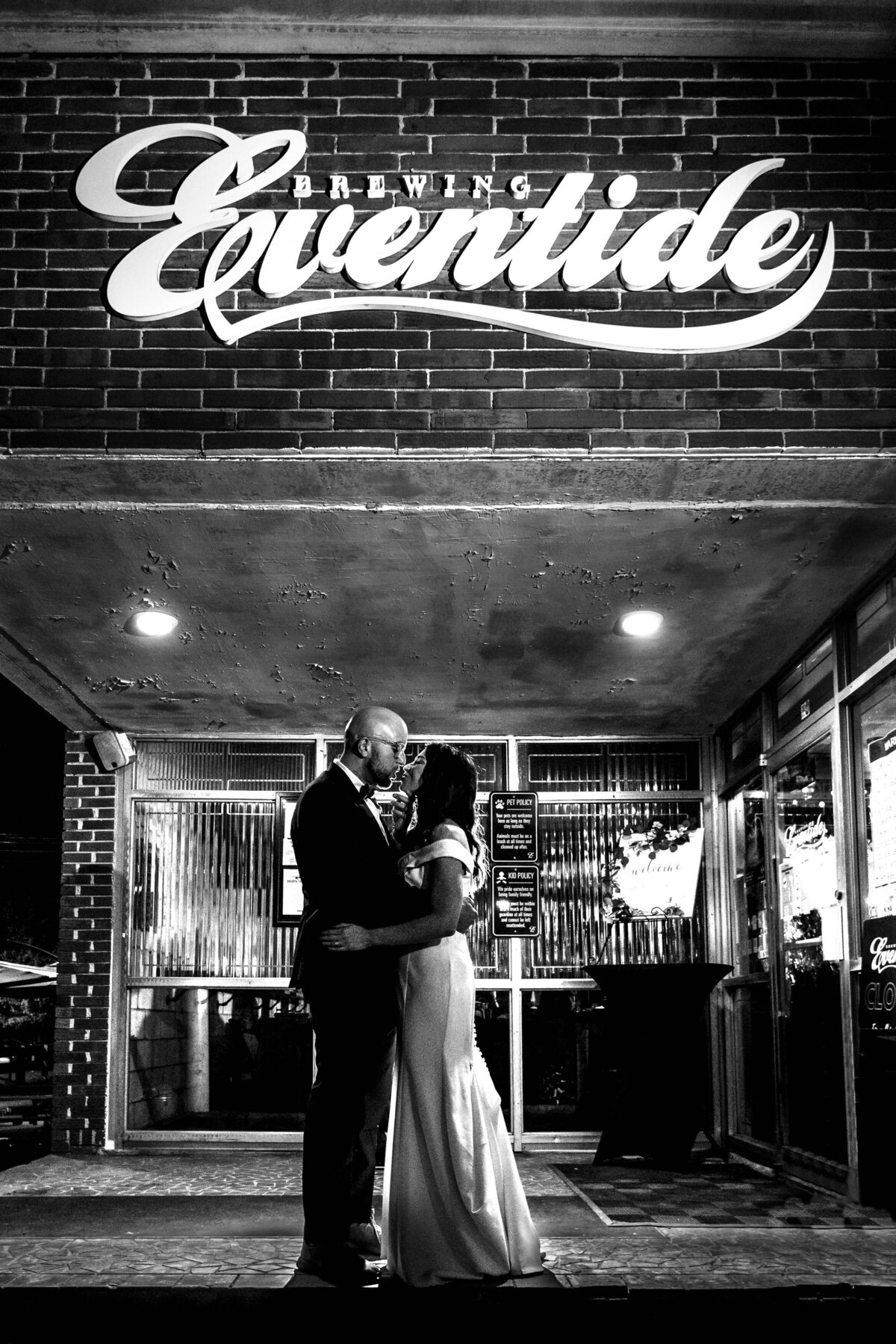 Black and white image of bride and groom with their arms around each others waists just before their lips meet for a kiss under the Eventide Brewing sign  in Atlanta by Charlotte wedding photographers DeLong Photography