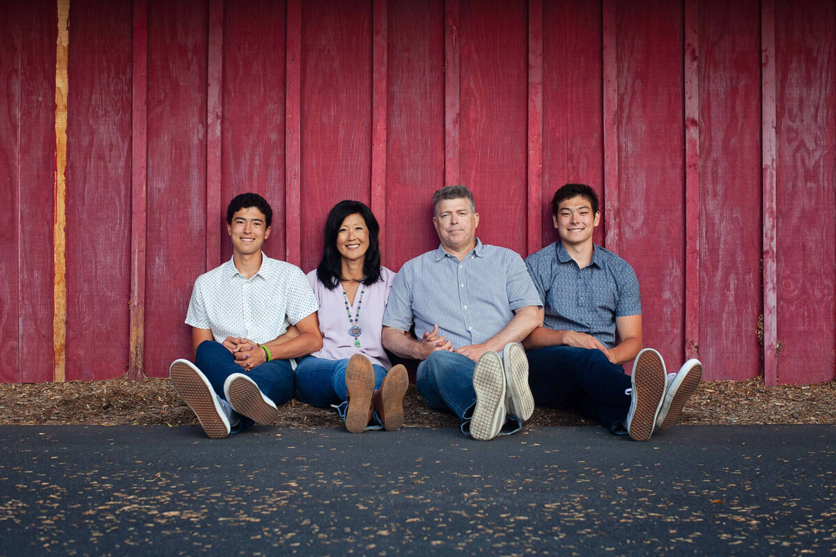 A family of four with dark hair sitting next to each other on asphalt with their feet crossed at the ankles and their backs against a red barn door at Irvine Ranch Park.