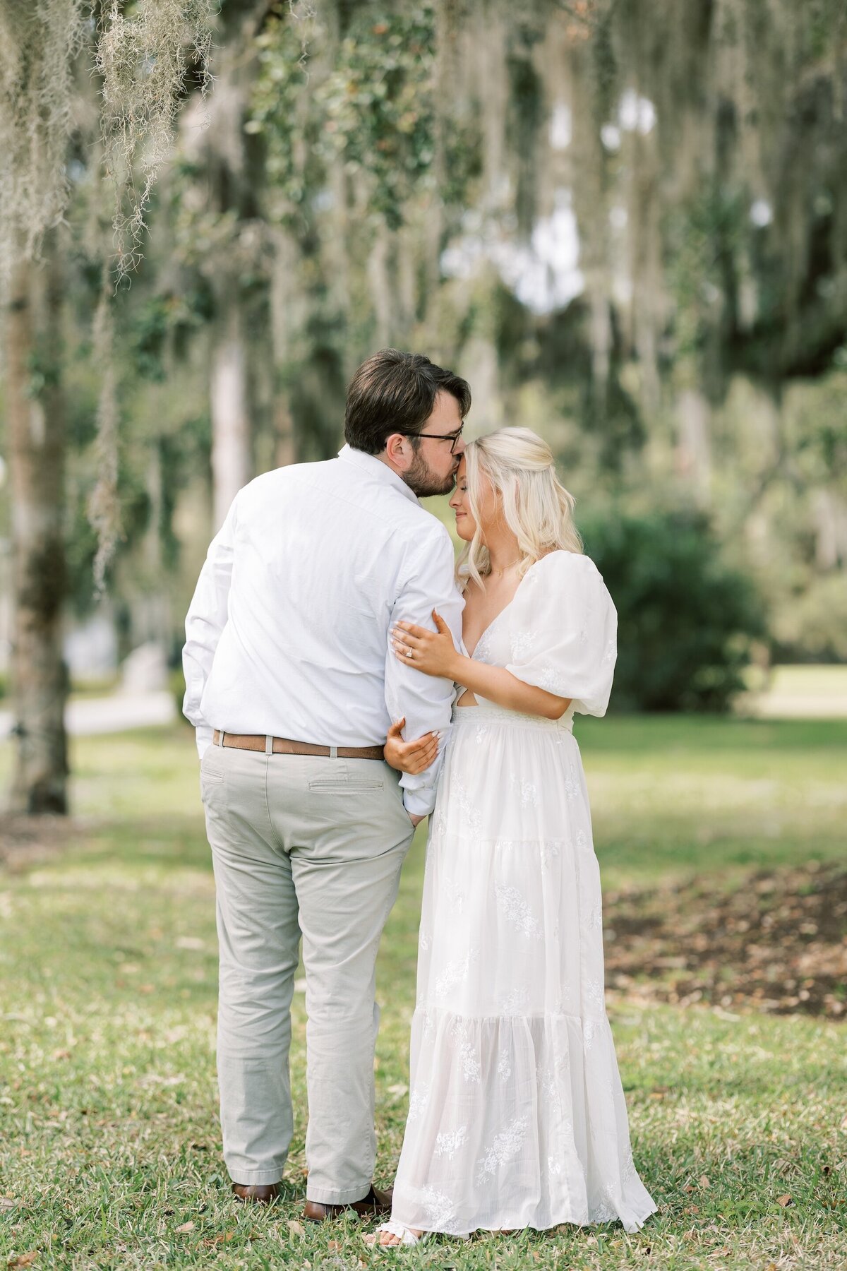 man kissing his fiance on the forehead as they stand under hanging moss trees