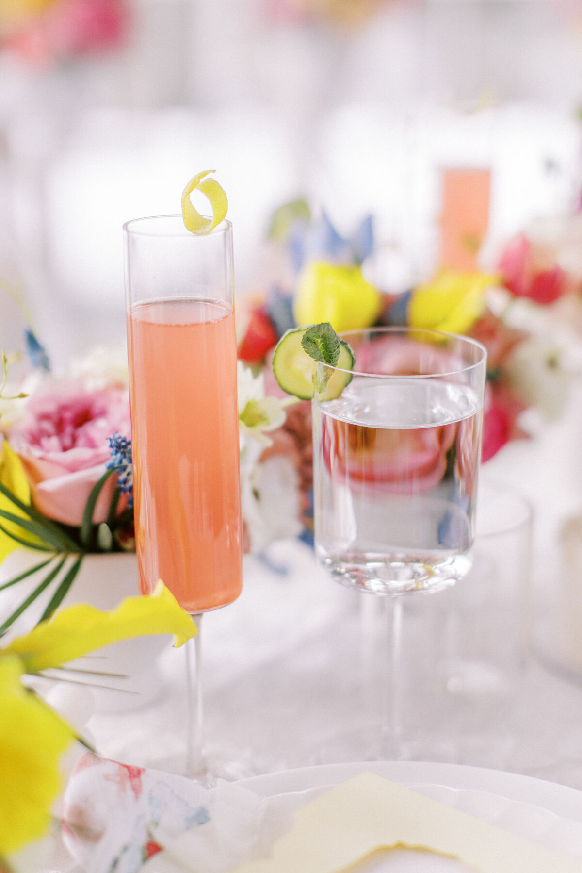 Two glasses on a table: one with a pink cocktail garnished with a lemon twist, the other with water and a lime slice. The table, perfect for weddings in Banff Alberta, is adorned with colorful flowers.