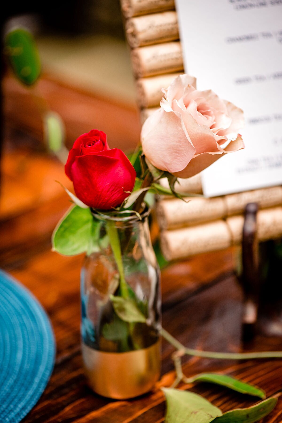 Red rose and blush rose in a glass bud vase with a gold base sitting on a dark wood farm table next to  a bar menu at a summer wedding at Arrington Vineyards.