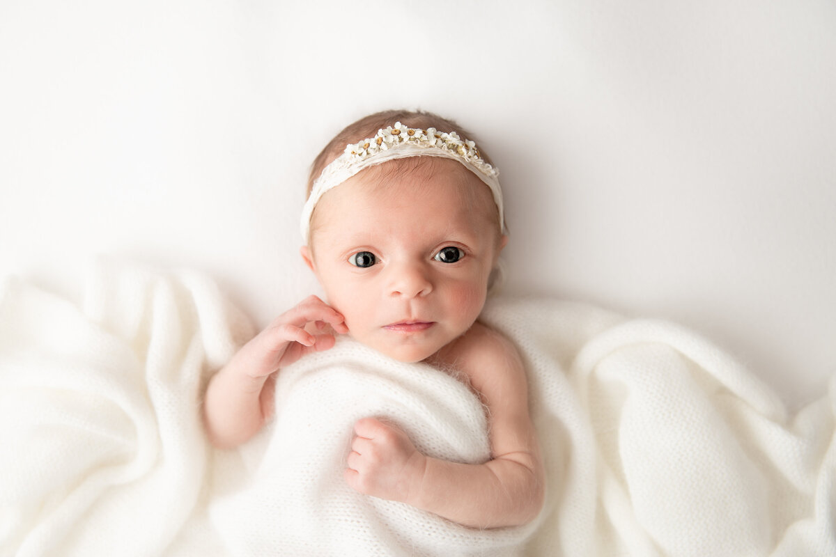 Newborn baby lying on white fabric with a white blanket over her looking directly at the camera.