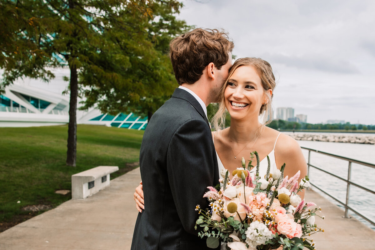 A bride laughs while being hugged by her groom on a waterfront walkway