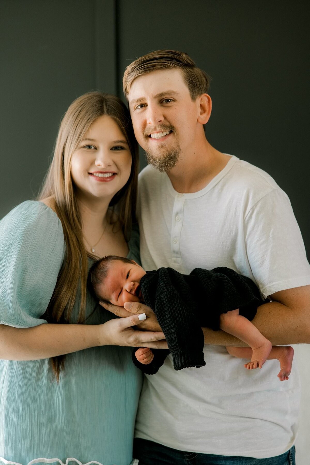 first time parents smiling at the camera, dad is holding baby on his forearm.