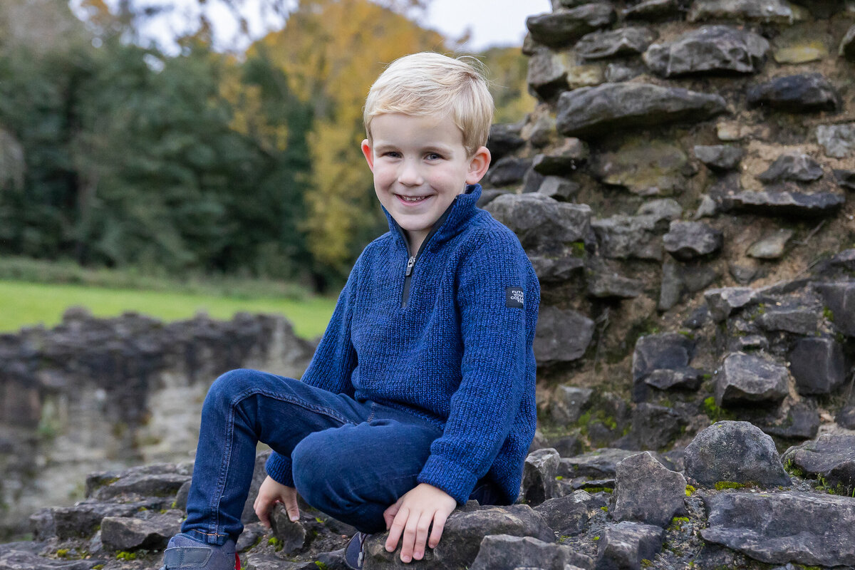 Young boy with blonde hair smiling and seated on a stone wall, wearing a blue sweater and jeans, with greenery in the background.