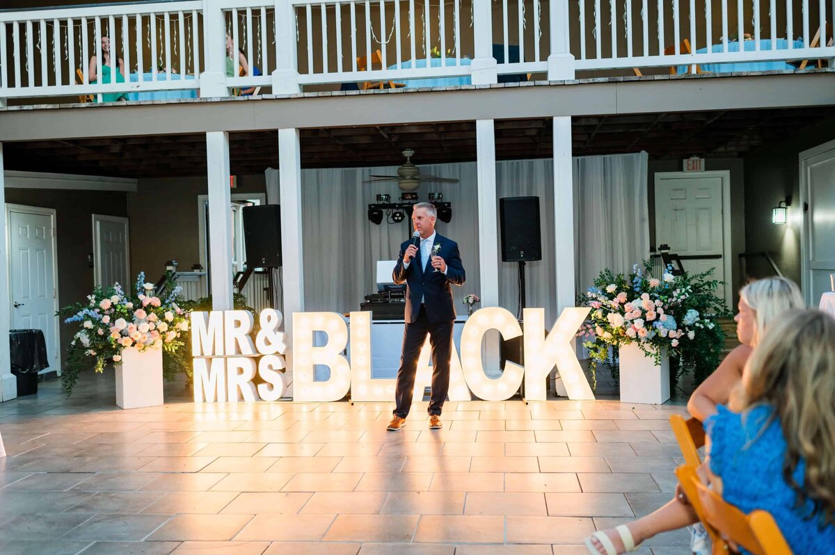 A man in a suit speaks into a microphone at an outdoor courtyard wedding. Behind him, large, illuminated letters spell out “MR and MRS.” Flowers decorate the area, and guests are seated at tables in the foreground.